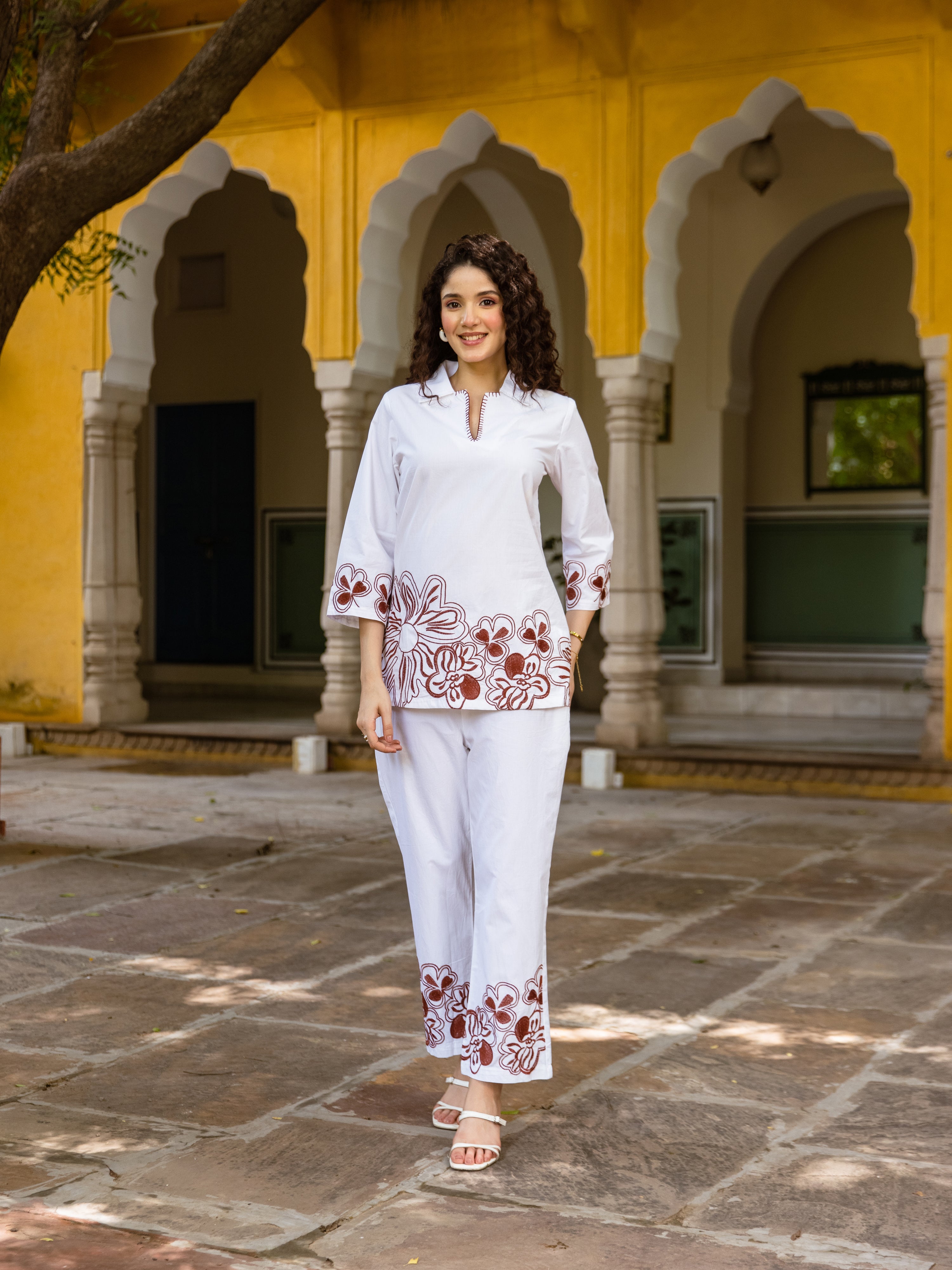 Woman wearing a white co-ord set with brown floral embroidery on the hem and sleeves, posing in front of a traditional Indian arched courtyard.